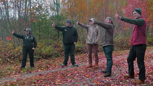 Five students standing in the woods in autumn, holding their thumbs out