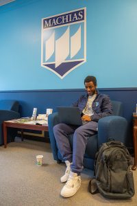 A student uses a laptop while sitting in a blue chair. The wall behind is dark blue on the bottom and light blue on the top. There is a Machias crest logo on the wall.