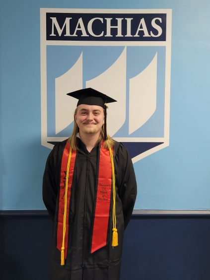 Evan has should length blonde hair. He is wearing a black graduation gown with a red sash and yellow honor cords. The background is blue and white