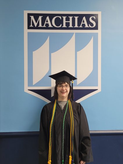 Keely has shoulder length brown hair and is wearing a black graduation gown with yellow honor cords . The background is blue and white