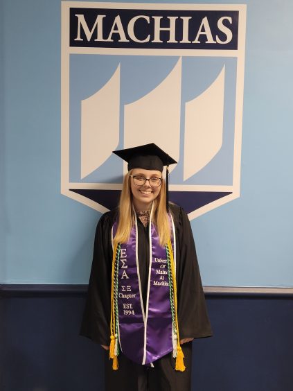 Rachael has long blonde hair. She is wearing a black graduation gown with a purple sash and yellow honor cords. The background is blue and white