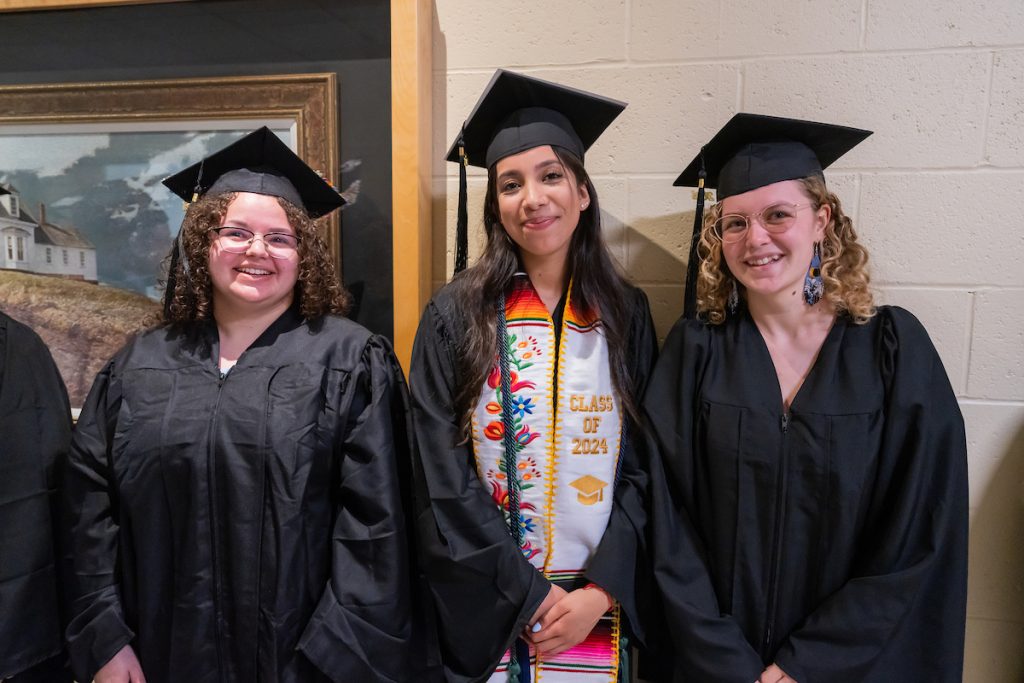 3 students pose together in the hallway in their black caps and gowns before marching into commencement. 