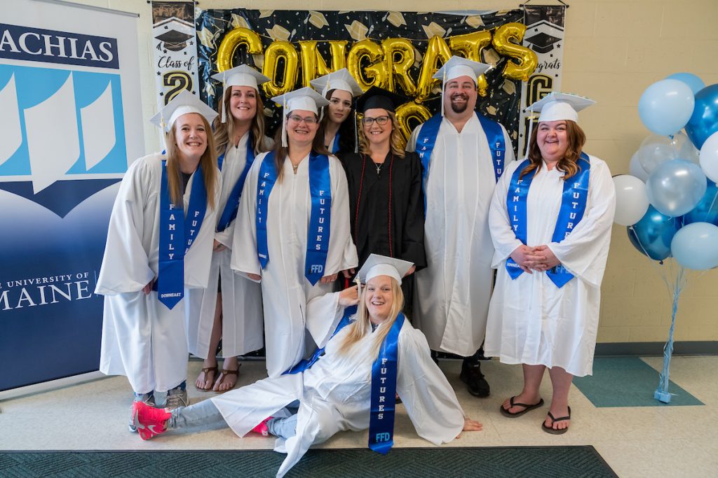 8 students pose before graduation in their caps and gowns in front of blue, white, black and gold decorations. 