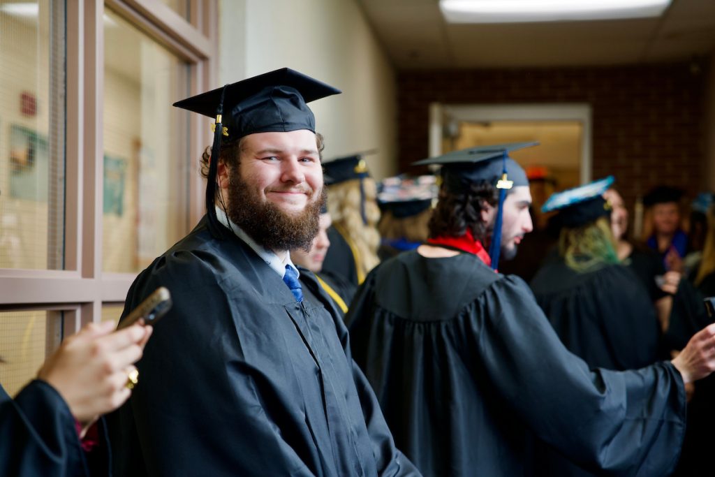 A student smiles at the camera in their cap and gown during lineup before commencement. Other students are talking behind them.