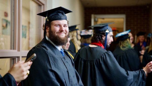 A student smiles at the camera in their cap and gown during lineup before commencement. Other students are talking behind them. 