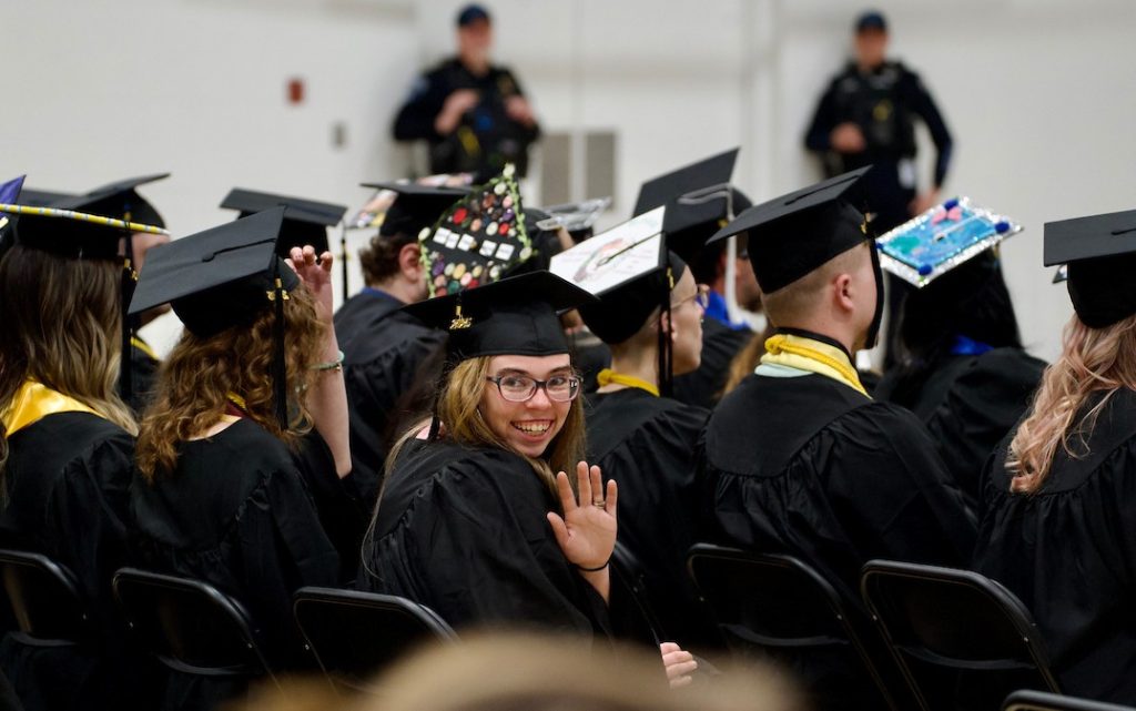 A graduate turns around and waves to their family while sitting with the rest of the graduating class in their caps and gowns.