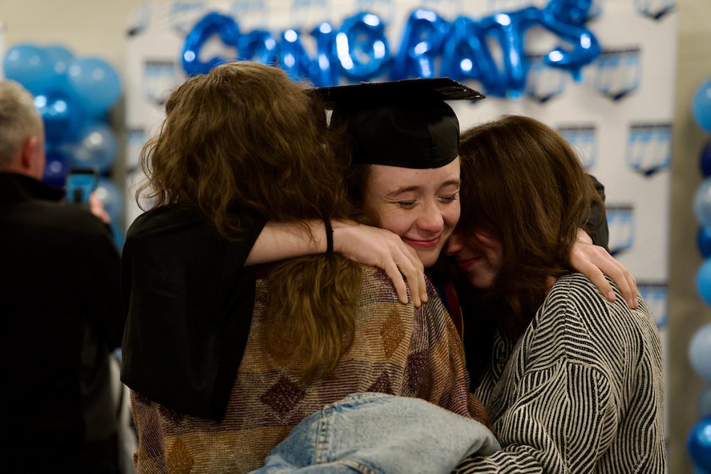 A graduate hugs two people after commencement. A balloon banner is behind them that says "Congrats"
