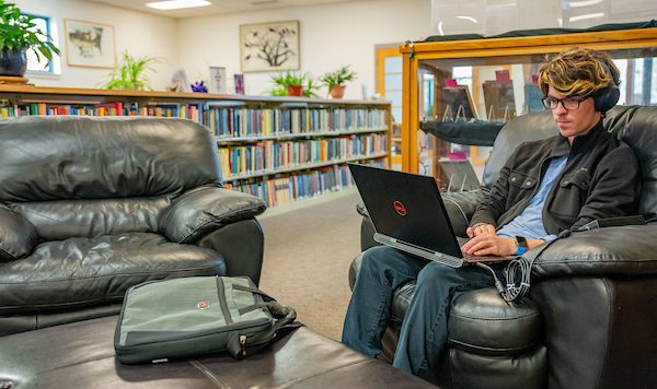 A student sits in a black leather chair in the library while using a laptop and wearing headphones. Shelves of books are in the background.