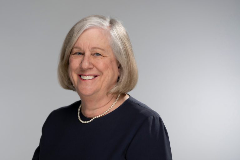 President Ferrini-Munday is wearing and black shirt and white necklace. The photo has a gray background.