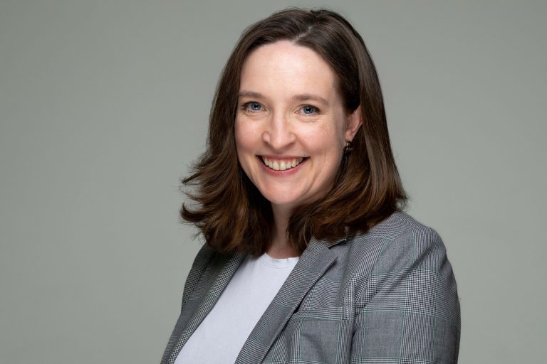 Dean Walsh has on a white shirt and gray blazer. The background is light gray.