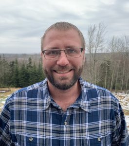 Kyle has very short hair, wears glasses and has a goatee. He is wearing a blue and white checkered button up shirt. The background is tree that have lost their leaves due to winter. 