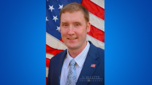 Philip has short light brown hair. He is wearing a dark blue suit, white and blue striped shirt and light blue tie. The american flag is the background.
