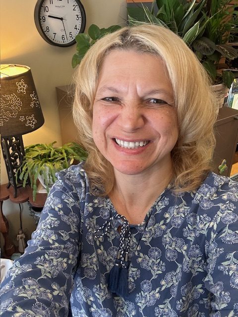 Stacey has shoulder length blonde hair and is wearing a blue shirt with white florals. 