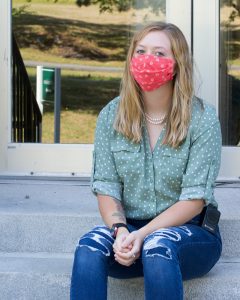 A female student is seated on concrete steps. She is wearing an orange cloth face mask