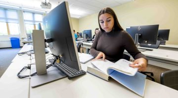 A student with long brown hair and wearing a long sleeve brown shirt, sits in a computer lab while looking in her textbook.