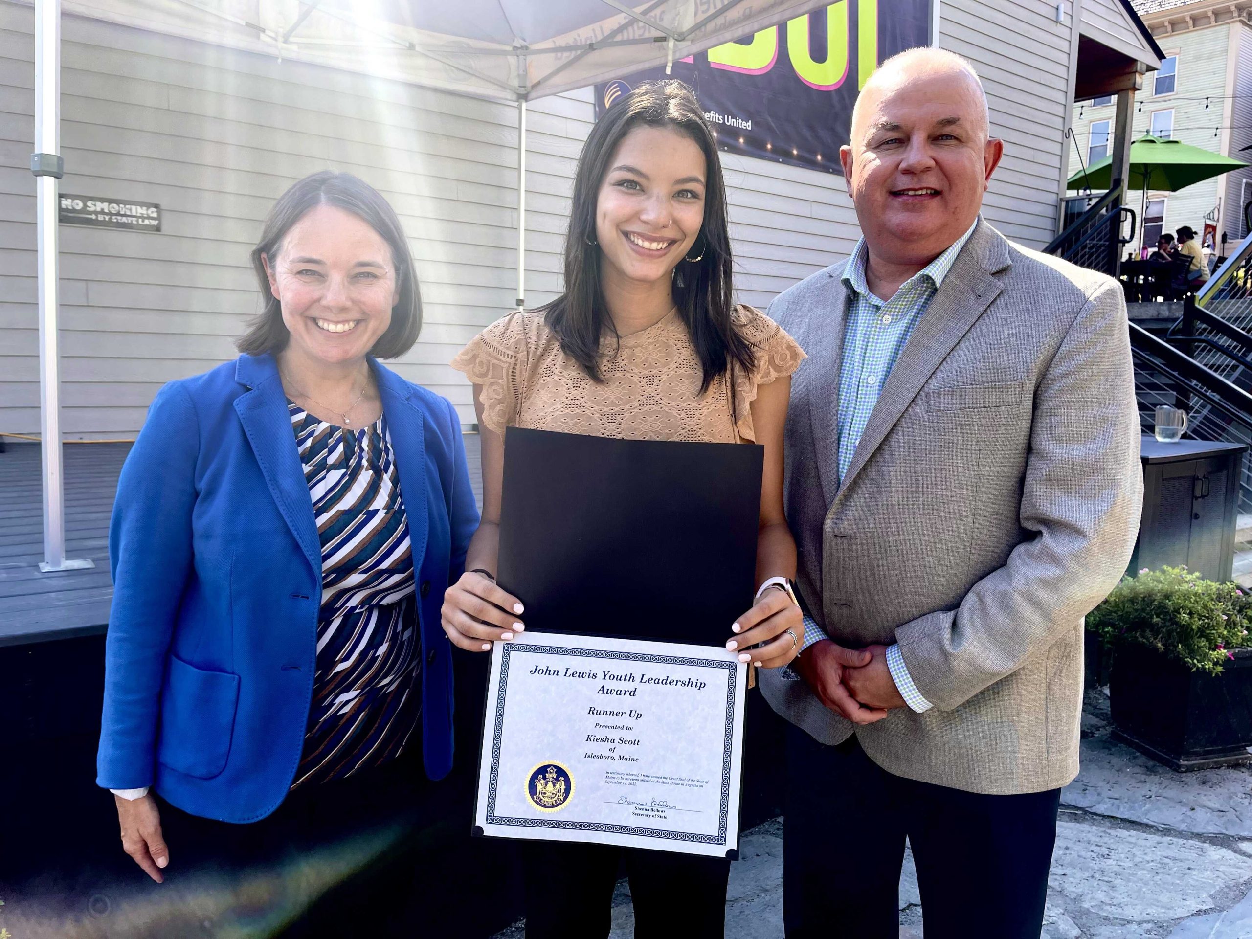 Maine Senator Shenna Bellows, UMaine Machias student Kiesha Scott and JMG CEO Craig Larrabee at the luncheon. They are standing outside in front of a building, Kiesha is holdnig her certificate.