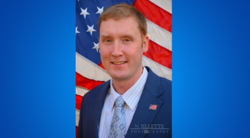 Philip has short light brown hair. He is wearing a dark blue suit, white and blue striped shirt and light blue tie. The american flag is the background.