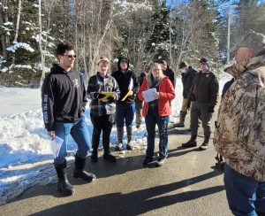 A handful of students stand to the left along with the professor and a game warden. They are facing a gentleman who stands to the right wearing a camouflage jacket