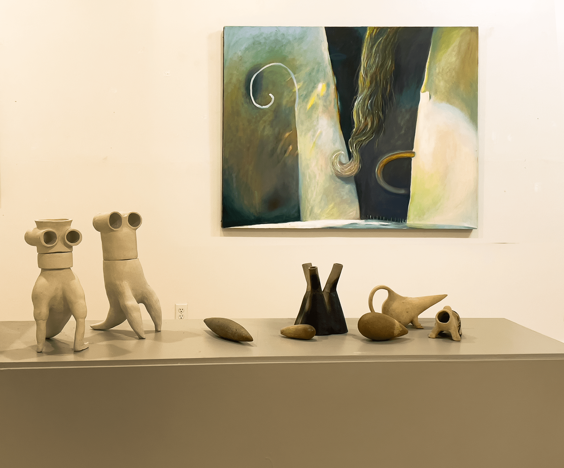 An abstract painting in greens and neutral tones hangs on a cream colored wall. A handful of pottery pieces are on a stand in the foreground.