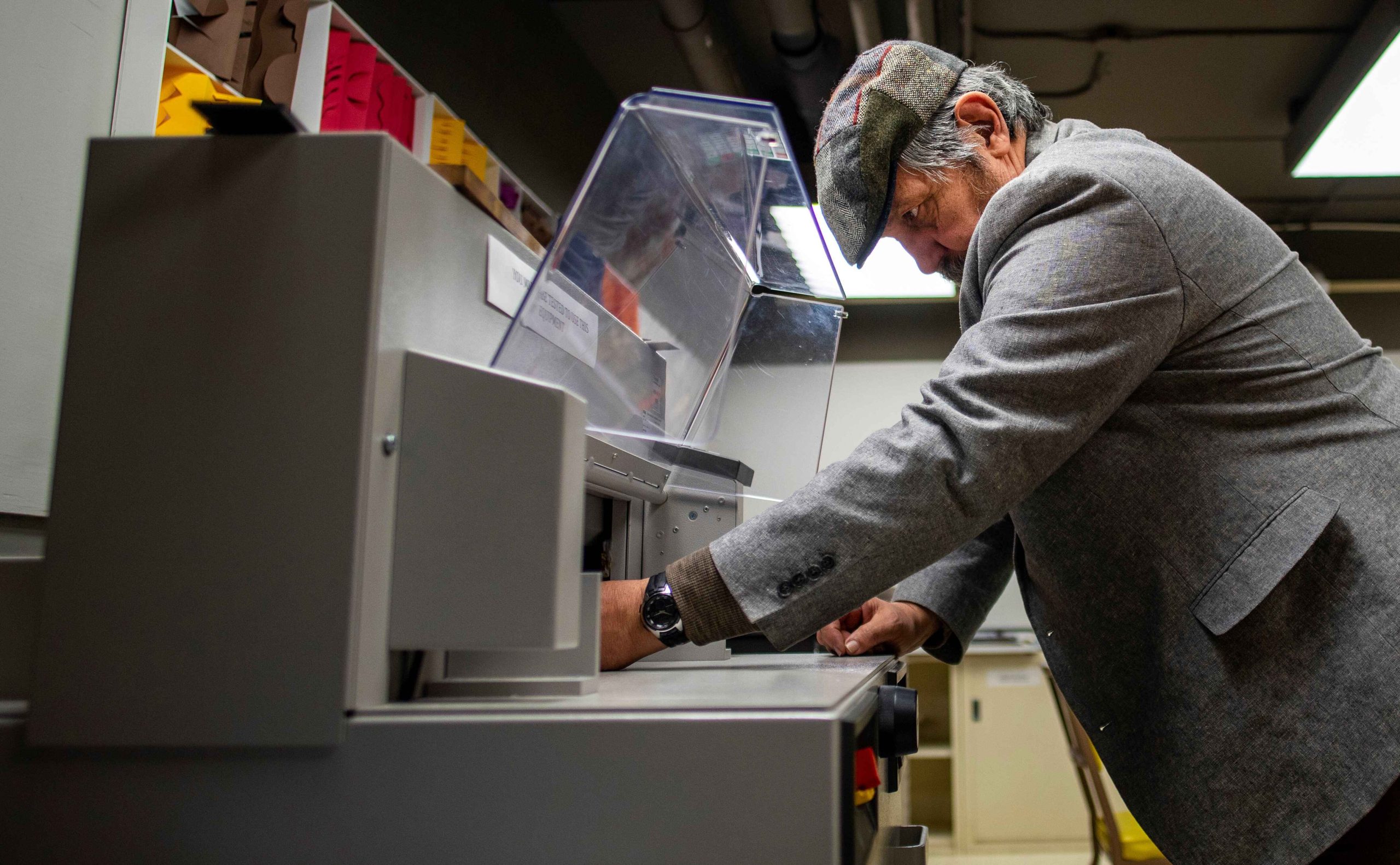 Bernie Vinzani, wearing a grey coat and cap, works with a machine in the book arts studio