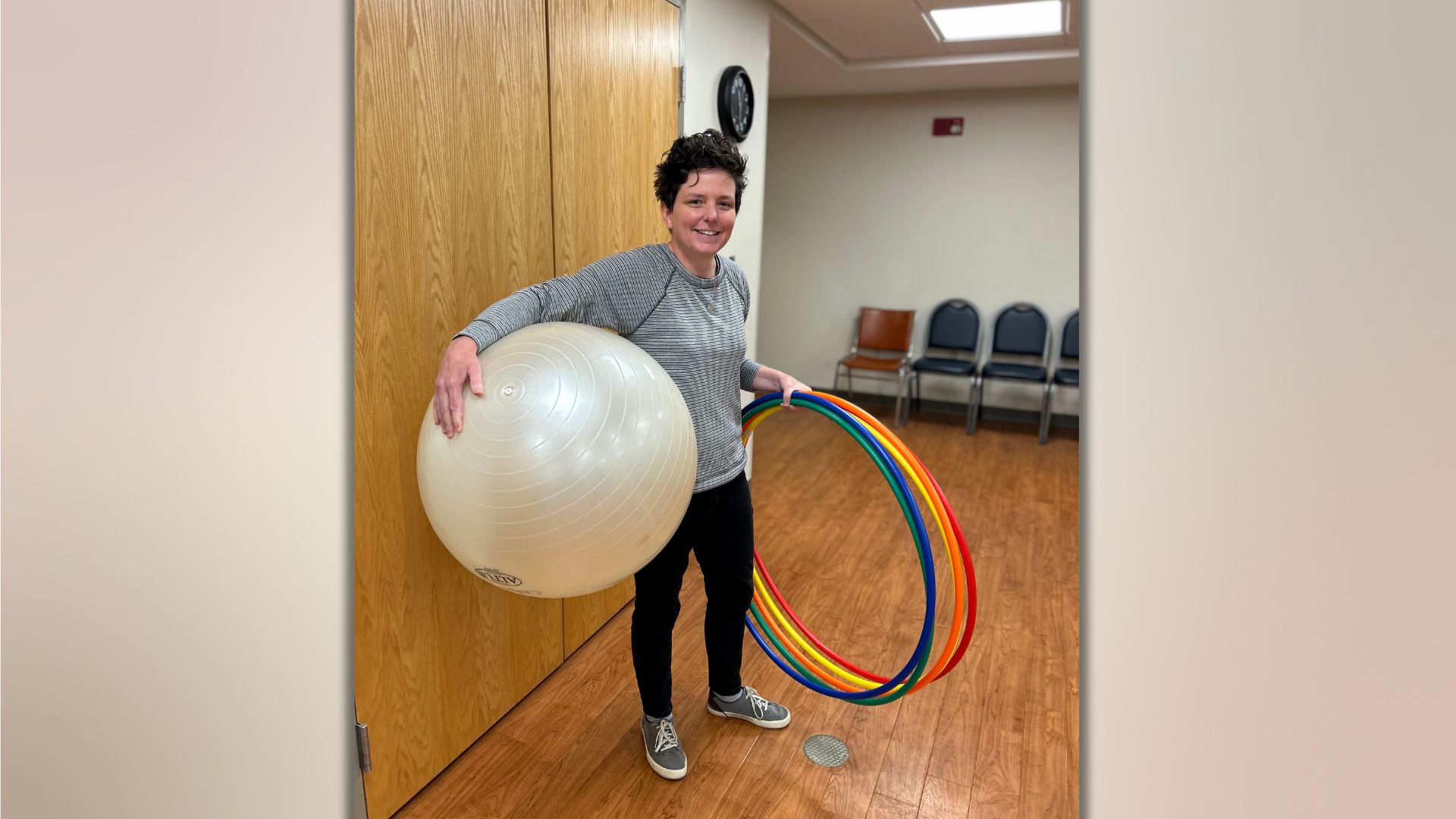 Melissa has short brown hair and the background consists of a wooden floor and wooden walls. Melissa is holding a number of multi colored hoolahoops and a white yoga ball.