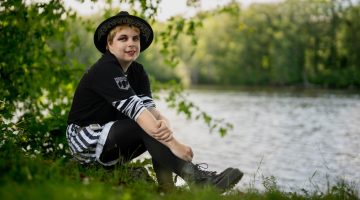 Xander poses by sitting on the bank of a local water way. He is wearing all black and white, including a black fedora and boots. .