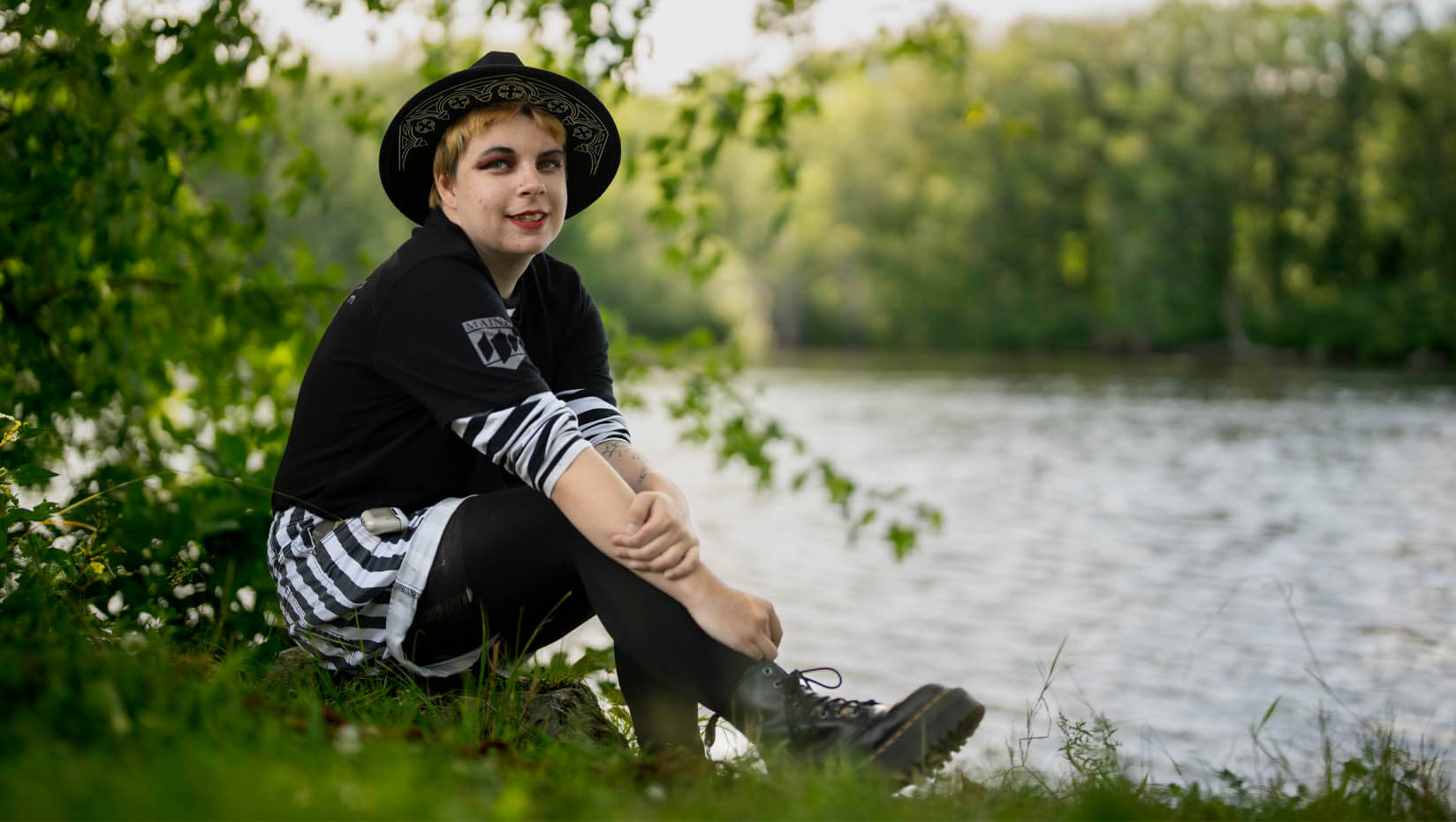 Xander poses by sitting on the bank of a local water way. He is wearing all black and white, including a black fedora and boots. .