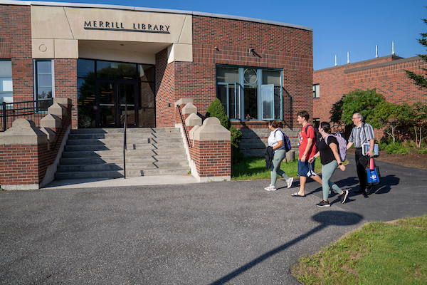 3 students and a professor walk in front of Merril Library. It's a sunny day. Merrill library is made of red bricks with concrete stairs.