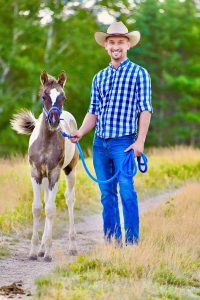 Nick wears jeans, a blue checkered shirt and western hat. He is outside standing next to his horse. 