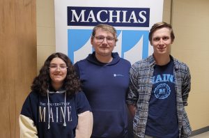 A photo of 3 students in front of a Machias banner. They are all wearing different UMaine and UMaine Machias shirts
