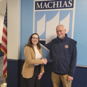 2 people shake hands. The UMaine Machias logo is in the background and an American flag to their left. 