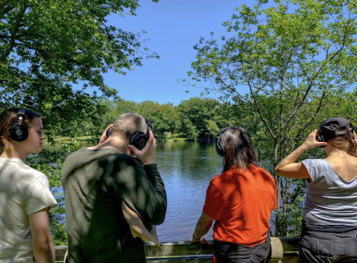 Four people stand by the water, listening to nature through headphones as they record.
