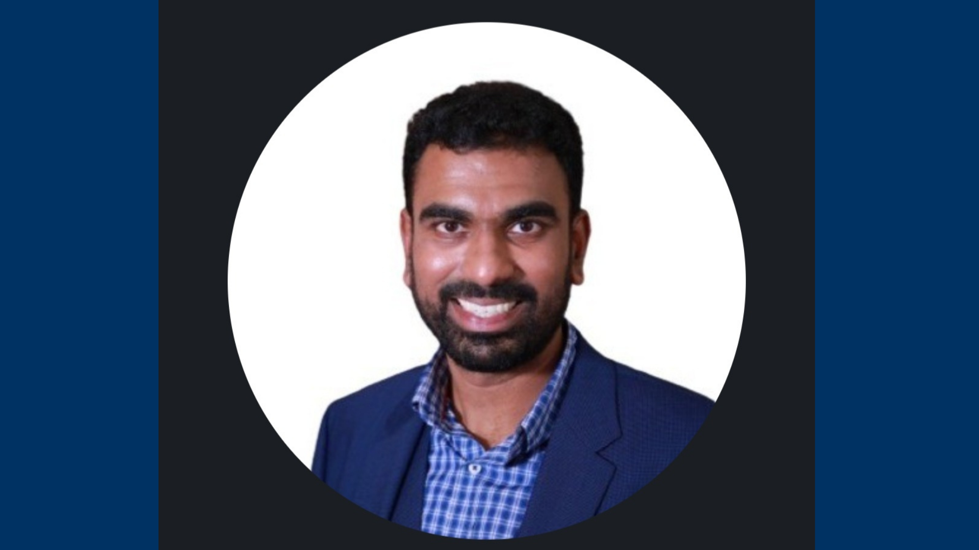 Vijay has short black hair. He is wearing a light blue and white checkered shirt and royal blue blazer.