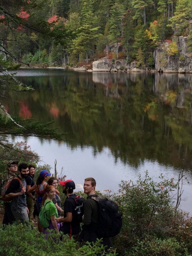 Students walk alongside a cliff, trees are shown reflected in a placid river