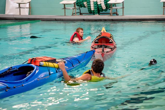 Indoor pool scene. Two individuals are next to their kayaks, wearing life jackets for a kayak rescue class.