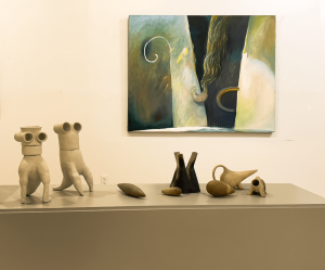 An abstract painting in greens and neutral tones hangs on a cream colored wall. A handful of pottery pieces are on a stand in the foreground.