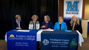 A panel of 5, including President Ferrini- Mundy of UMaine and UMaine Machias as well as President Susan Mingo of WCCC, sit behind a table with some banners on it.