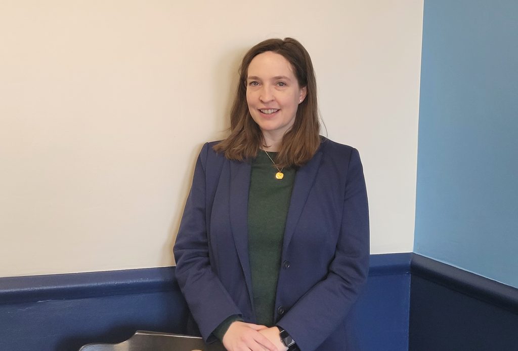 Megan poses in front of a white wall that is split with a dark blue bottom half. Her hands are on the back of a chair and she is wearing a blue blazer.