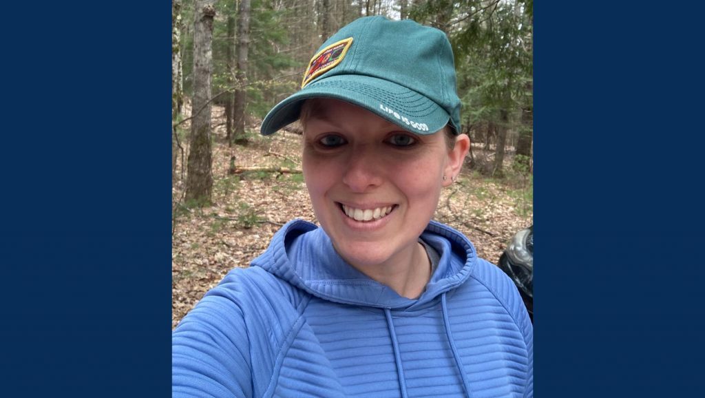 Kait is wearing a blue hooded sweatshirt and a light green hat. She is in a wooded area.