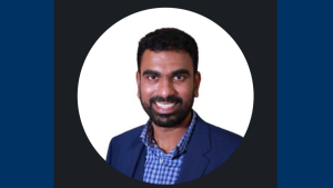 Vijay has short black hair. He is wearing a light blue and white checkered shirt and royal blue blazer.