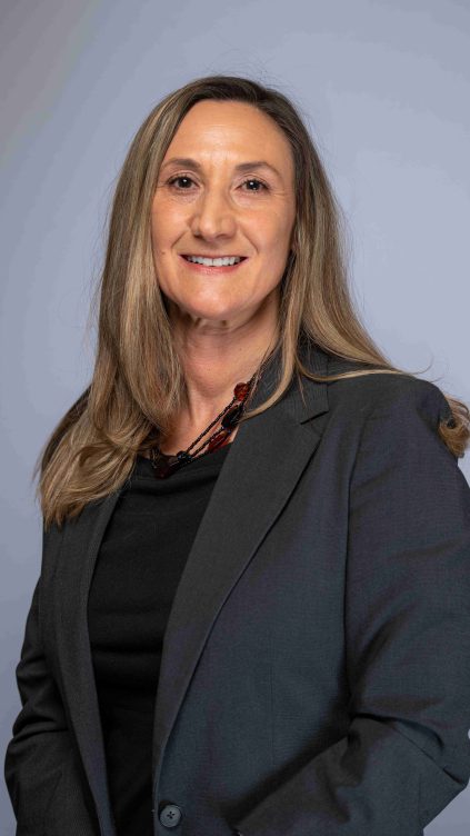 Heather has long blond hair and is wearing a black shirt and gray blazer in front of a blue and gray background