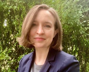 Megan Walsh stands in front of greenery.  She has sholder length brown hair and is wearing a gray shirt and dark blue blazer. 