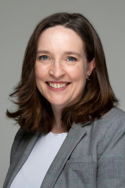 Dean Walsh has shoulder length dark hair and is wearing a white shirt and gray blazer.