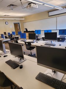 This computer lab has desk top computers set on tables of 3 rows. The rows are are split down the middle from the front to the back of the classroom for row access. The front of the room has a whiteboard and projector screen.