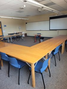 wooden tables are set up in a horseshoe patter in this classroom. The chairs are blue. There is a white board and a projector sreen at the front of the room.