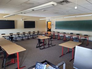 A classroom is set up with individually placed desks in a horseshoe pattern. There is a chalkboard on the right and tvs on the back wall.