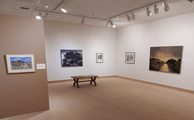 Exhibits from our Permanent Collection hung on the wall and spotlit from above. White and tan walls and a tan floor.