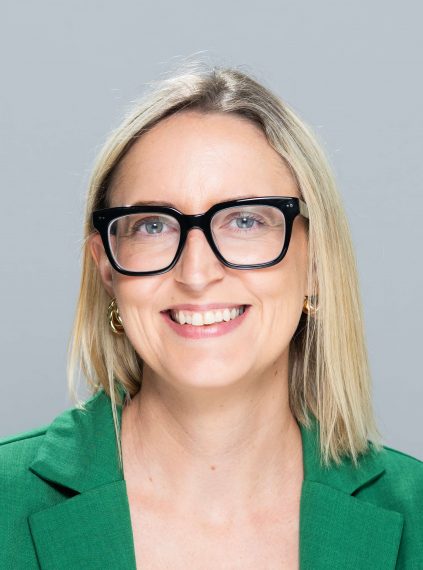 Danielle has shoulder length blond hair. She is wearing a green button up shirt and black glasses.